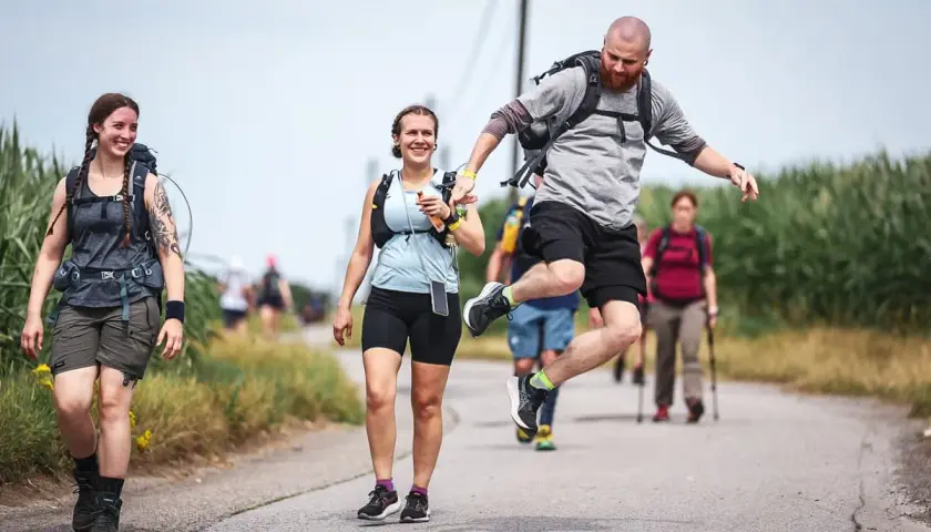 Wanderer mit Rucksäcken auf einem Feldweg, einer macht einen Sprung. Sie tragen bequeme Wanderschuhe, die man von bekannten Schuhmarken kennt.