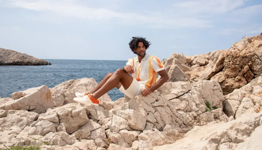 Mann sitzt auf Felsen am Meer, trägt ein buntes Poloshirt, weiße kurze Hose und passende schuhe zu kurzen hosen herren mit orangen Akzenten.