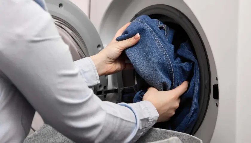 Hände legen Jeans in die Waschmaschine, um sie mit anderen Kleidungsstücken zu waschen.
