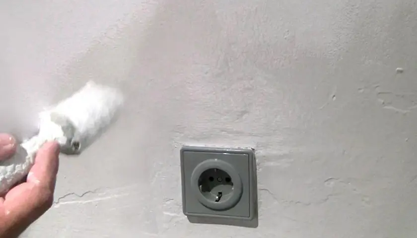 Hand mit Rolle tupft auf weiße Wand, um Wasserflecken zu entfernen. Steckdose im Bild.
