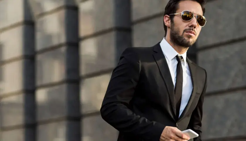 Ein Mann im Anzug mit Sonnenbrille hält ein Smartphone. Er steht vor einer modernen Gebäudefassade.