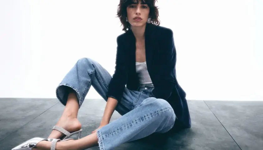 Frau sitzt lässig in Jeans und Blazer, zeigt, wie man Jeans elegant kombinieren kann. Glitzernde Schuhe runden den Look ab.
