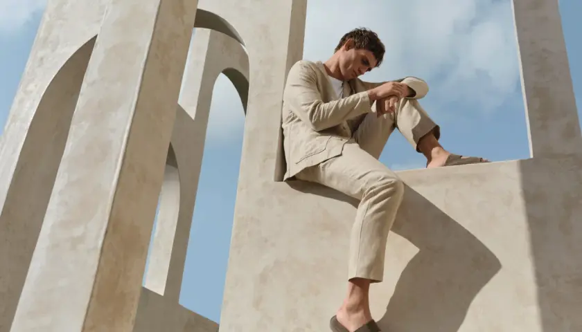 Mann in einem hellen, beigen Anzug mit einem blauen Sakko und einer beigen Hose sitzt auf einer Betonstruktur vor blauem Himmel.