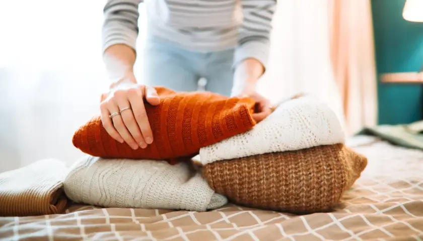 Hände falten einen Stapel warmer Strickpullover. Der orangefarbene Pullover liegt oben. Das Waschen von Wollpullovern erfordert Sorgfalt.