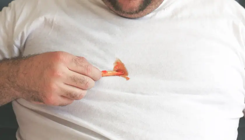 Mann mit fettigen Pommes auf weißem T-Shirt. Tipps, wie man fettflecken aus kleidung entfernen nach dem waschen kann.