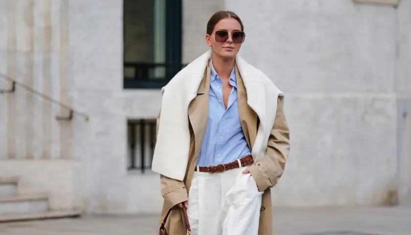 Eine Frau trägt einen beigefarbenen Trenchcoat, ein hellblaues Hemd und weiße Hose. Das Outfit zeigt, wie man ein beiges Oberteil kombinieren kann, auch für Herren.