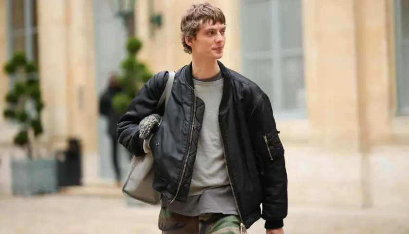 Ein Mann trägt eine schwarze Bomberjacke, die er lässig mit einem grauen Pullover und Camouflage-Hose kombiniert. Perfekt für den lässigen Herren-Look.