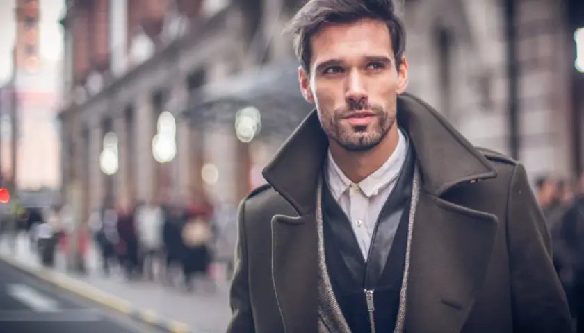 Ein Mann im eleganten Trenchcoat Herren Style, mit Bart und dunklem Haar, blickt nachdenklich in die Ferne.