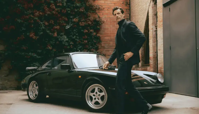 Ein Mann im eleganten Anzug mit Stiefel lehnt sich an einen klassischen Porsche.