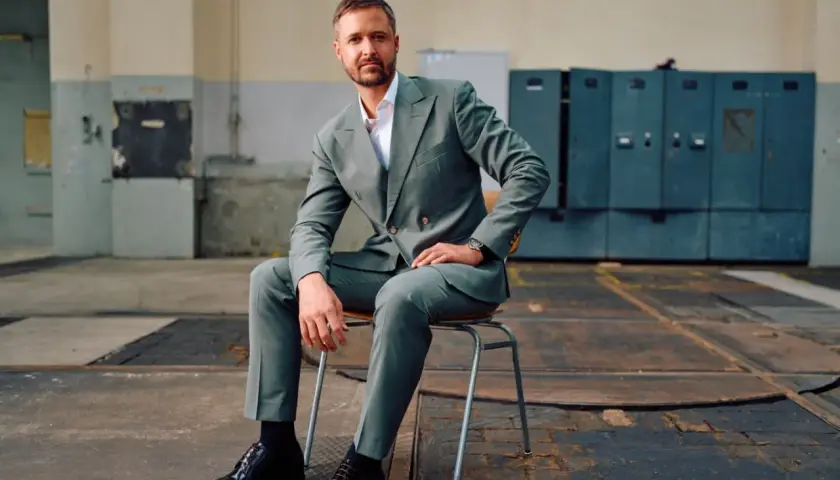 Mann in grauem Anzug mit Sakko mit 2 Schlitzen hinten, sitzt auf einem Stuhl in einer Industriehalle.