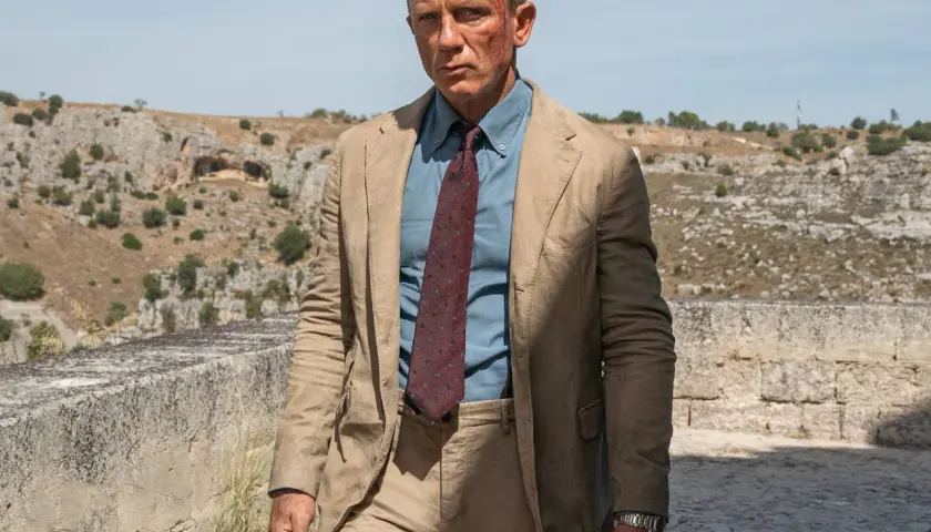 Daniel Craig in einem hellbraunen Anzug, einem blauen Hemd und einer gemusterten Krawatte, typisch für James Bond Outfits, mit leichten Verletzungen im Gesicht.