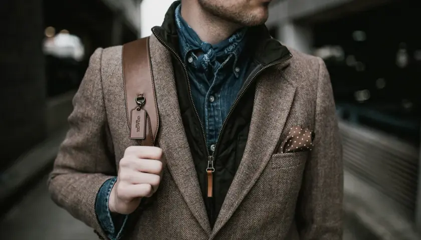 Mann im Tweed-Sakko mit Jeanshemd und Bandana, perfekt für den dresscode chic herren. Braune Ledertasche mit Coach-Anhänger.