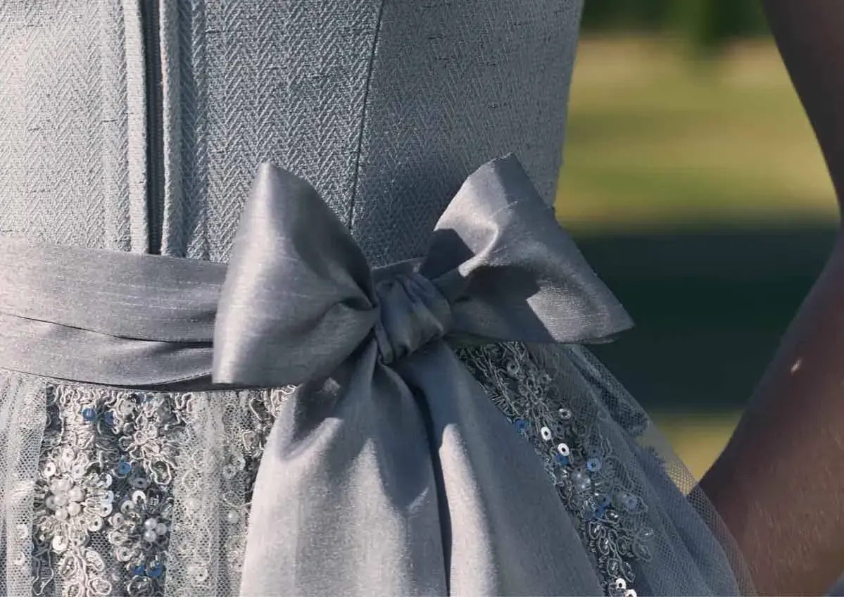 Ein Dirndl mit einer großen, grauen Schleife. Die Schleife ist Teil des Kleides, das auch mit Pailletten und Stickereien verziert ist. Perfekt für Kinder, die sich festlich kleiden möchten.