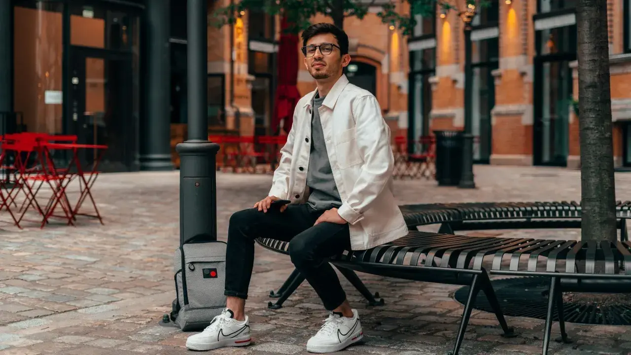 Ein Mann im smart casual Look für den Sommer sitzt auf einer Bank. Er trägt eine weiße Jacke über einem grauen T-Shirt, schwarze Jeans und weiße Sneaker.