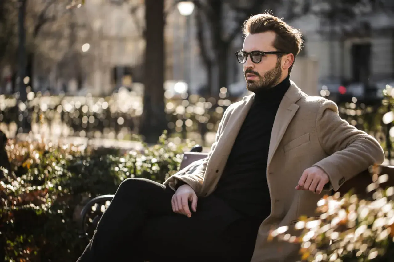 Ein Mann mit Bart und Brille sitzt stilvoll gekleidet in einem Park. Er trägt einen beigen Mantel über einem schwarzen Rollkragenpullover und dunklen Hosen.