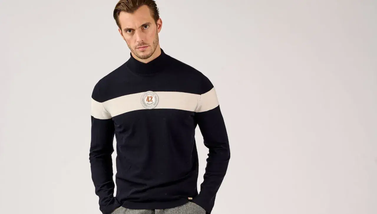 Mann in dunkelblauem Rollkragenpullover mit breitem cremefarbenem Streifen und Emblem. So kann man Rollkragenpullover kombinieren, Herren-Style.