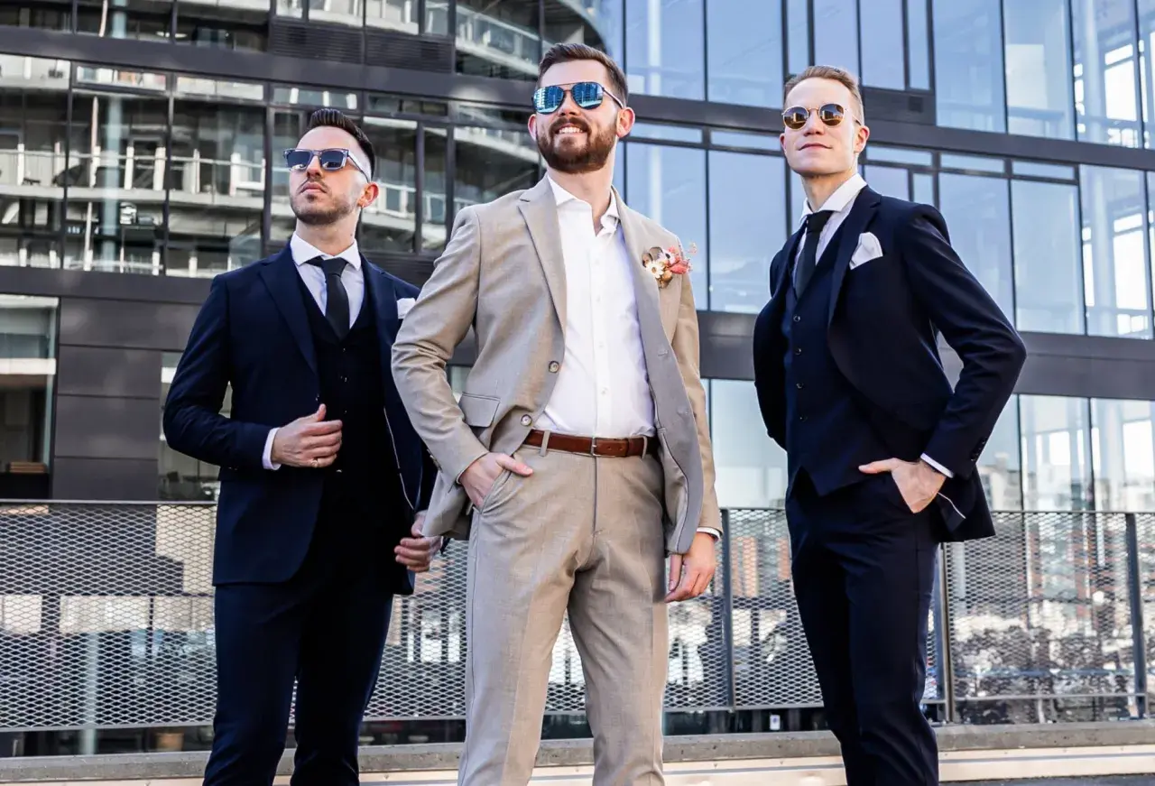 Drei Männer in Anzügen vor moderner Architektur. Der mittlere Mann trägt einen hellbraunen Anzug, die anderen dunkle. Eine stilvolle Inspiration, wie man schwarze Hose kombinieren kann für eine Hochzeit.