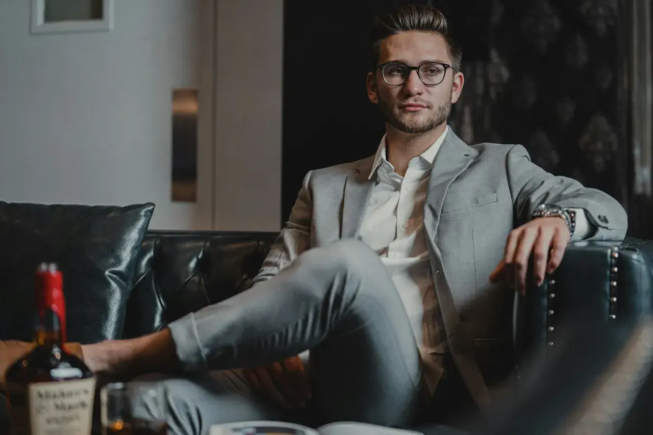Junger Mann in stilvoller Business-Kleidung für Herren, entspannt auf einem Ledersofa mit einem Glas Whiskey.