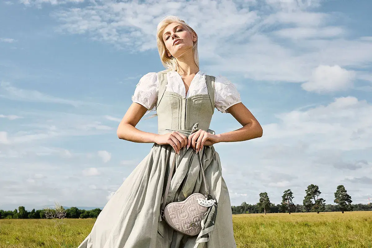 Eine Frau im Dirndl mit zarten Dirndl Farben steht auf einer grünen Wiese. Sie hält eine herzförmige Tasche.