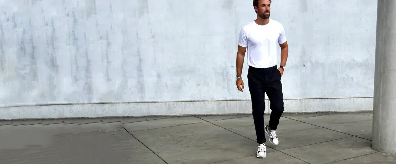 Mann in weißem T-Shirt und schwarzer Chino-Hose, dazu weiße Sneaker. Ein Beispiel, welche Schuhe zur Chino Hose Herren passen.