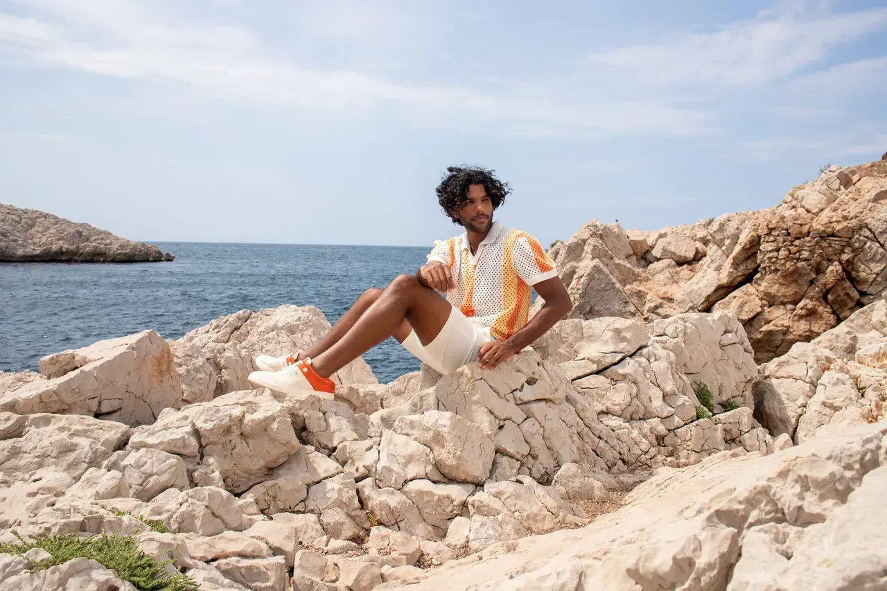 Mann sitzt auf Felsen am Meer, trägt ein buntes Poloshirt, weiße kurze Hose und passende schuhe zu kurzen hosen herren mit orangen Akzenten.