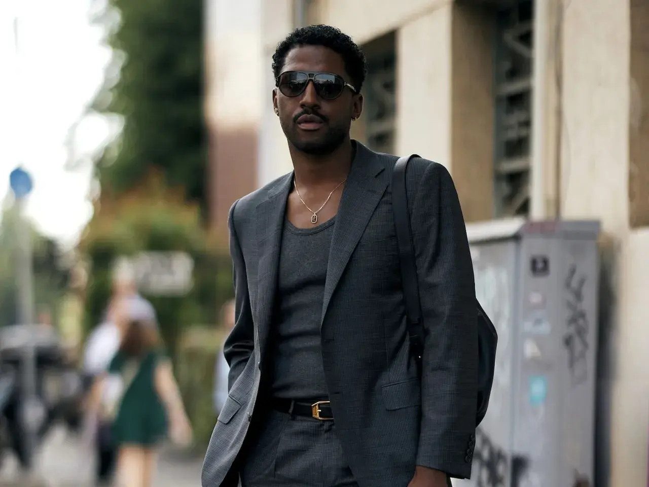 Ein Mann trägt einen eleganten Anzug mit Sneaker, dazu eine Kette und Sonnenbrille.