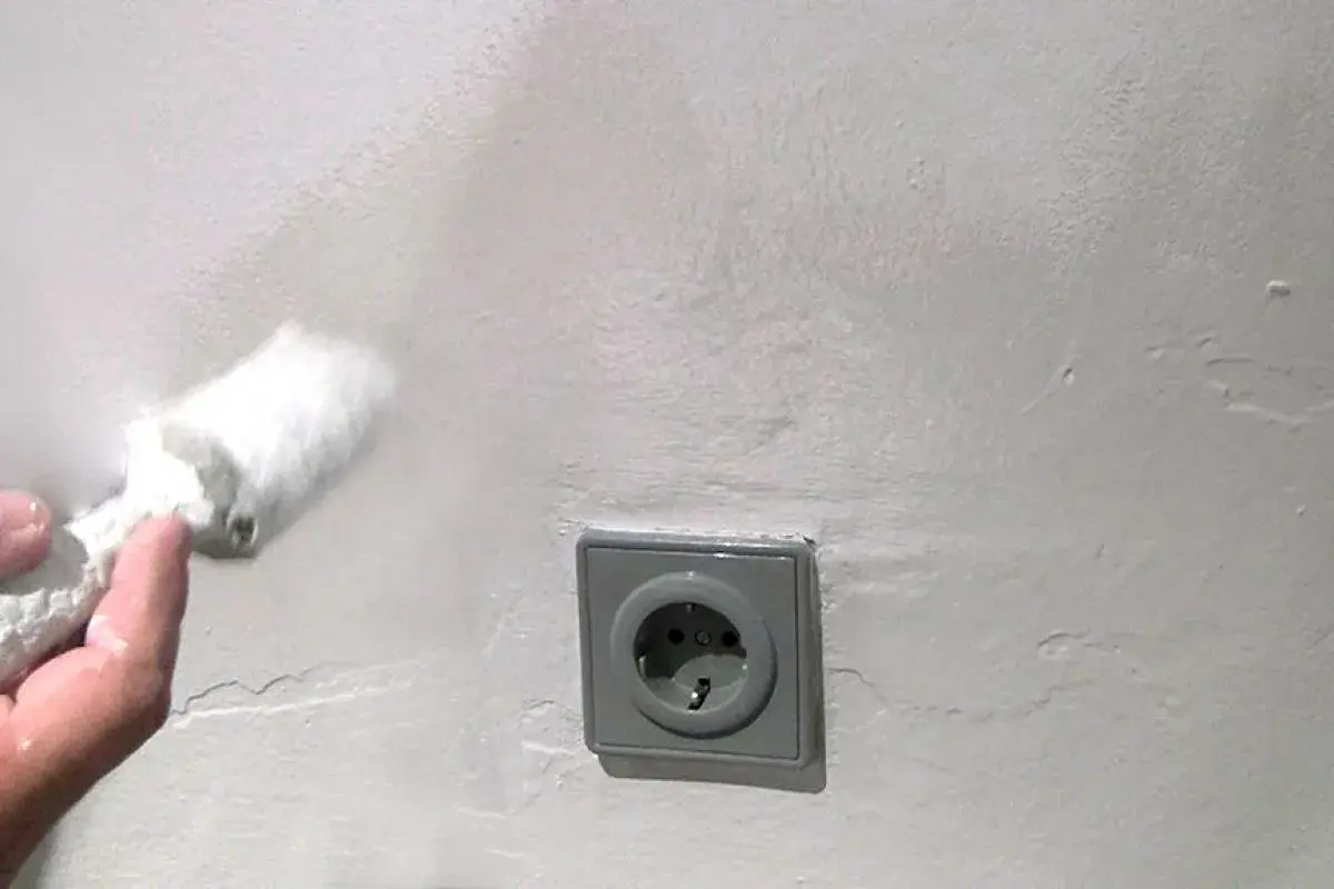 Hand mit Rolle tupft auf weiße Wand, um Wasserflecken zu entfernen. Steckdose im Bild.
