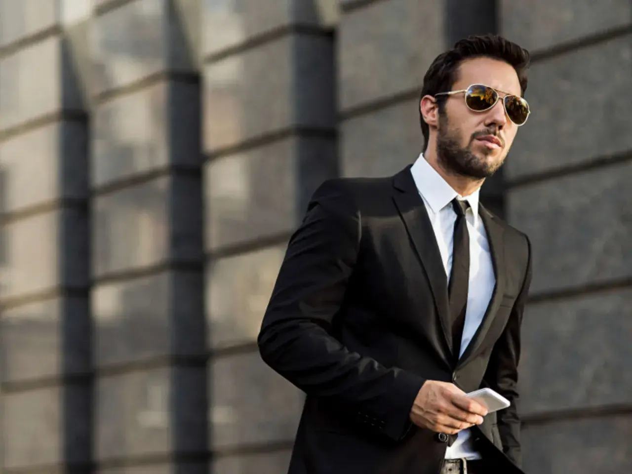 Ein Mann im Anzug mit Sonnenbrille hält ein Smartphone. Er steht vor einer modernen Gebäudefassade.