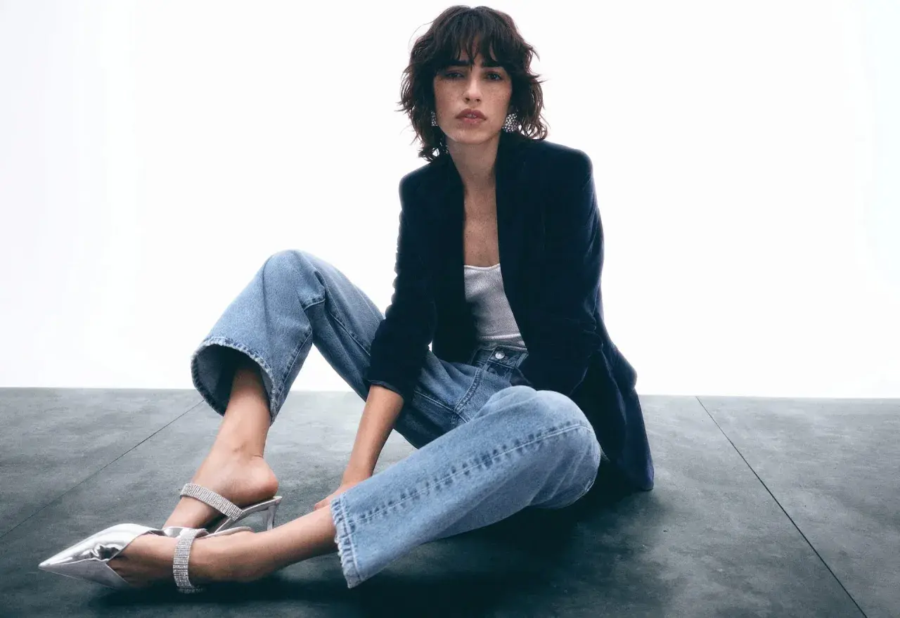 Frau sitzt lässig in Jeans und Blazer, zeigt, wie man Jeans elegant kombinieren kann. Glitzernde Schuhe runden den Look ab.