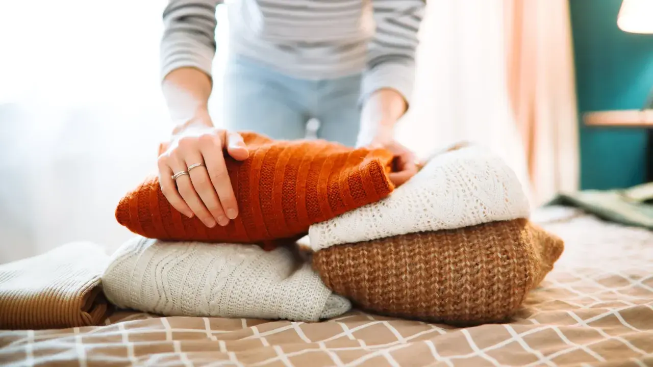 Hände falten einen Stapel warmer Strickpullover. Der orangefarbene Pullover liegt oben. Das Waschen von Wollpullovern erfordert Sorgfalt.
