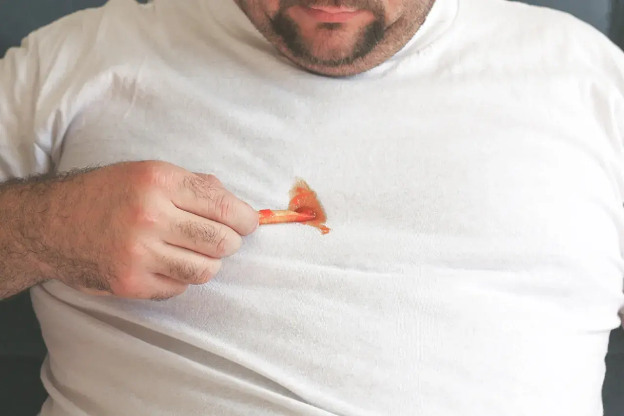 Mann mit fettigen Pommes auf weißem T-Shirt. Tipps, wie man fettflecken aus kleidung entfernen nach dem waschen kann.