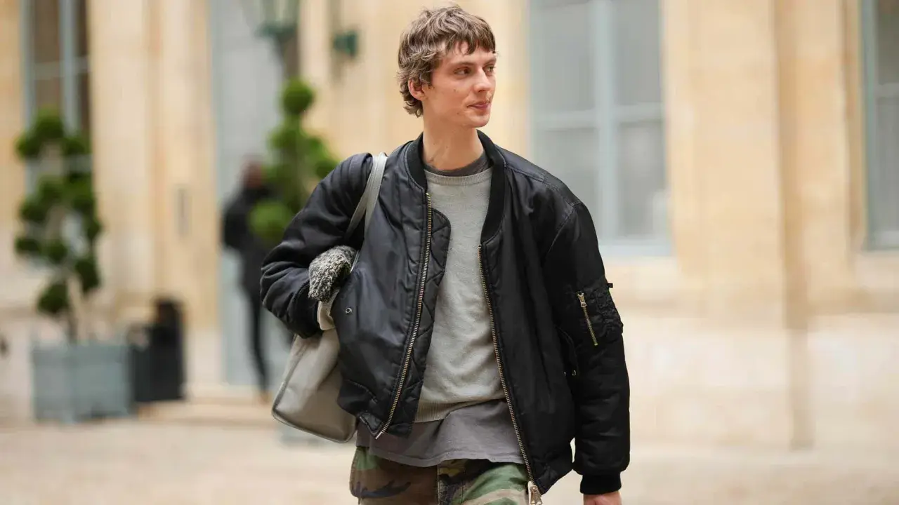 Ein Mann trägt eine schwarze Bomberjacke, die er lässig mit einem grauen Pullover und Camouflage-Hose kombiniert. Perfekt für den lässigen Herren-Look.