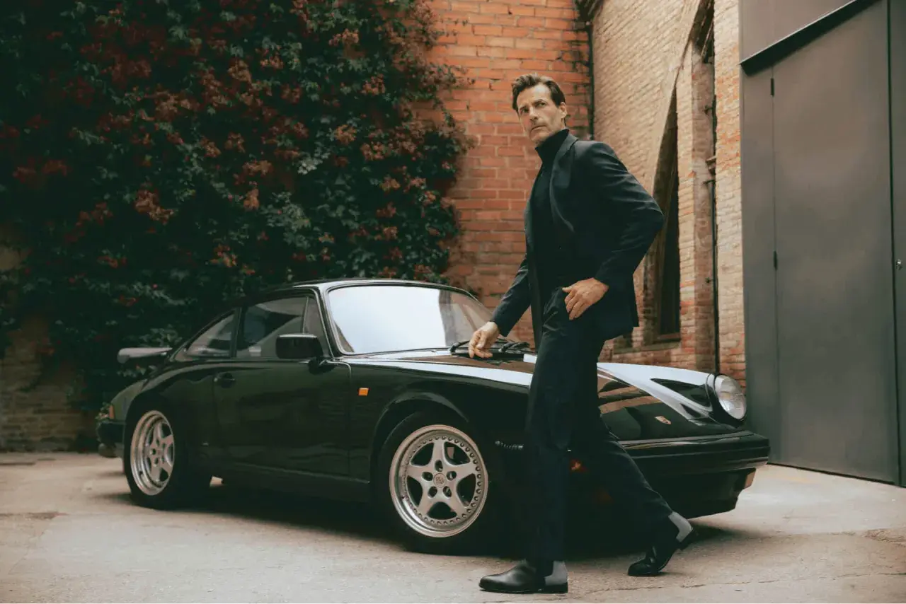 Ein Mann im eleganten Anzug mit Stiefel lehnt sich an einen klassischen Porsche.