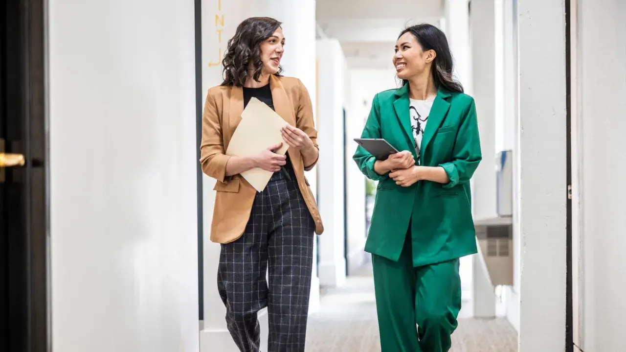 Zwei Frauen in **business casual** Outfits gehen lächelnd einen Flur entlang. Eine trägt einen braunen Blazer, die andere einen grünen Hosenanzug.