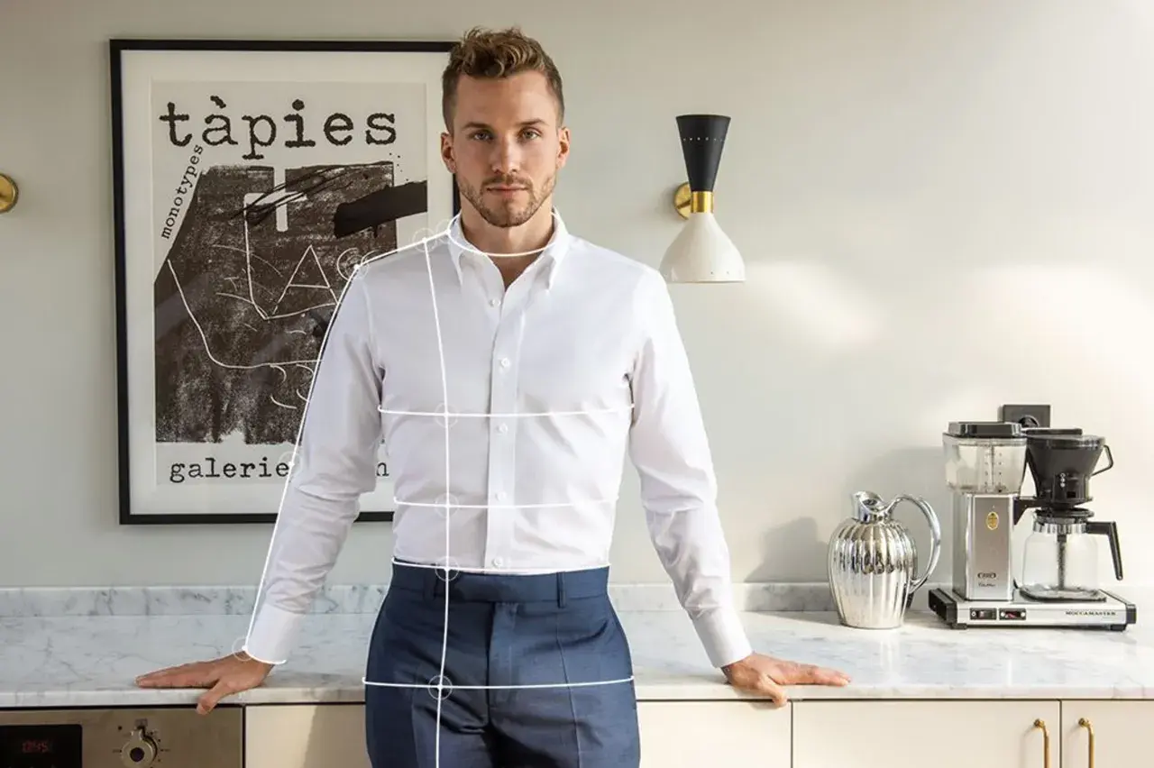 Mann mit weißem Hemd und blauer Hose, bereit, die Ärmel hochzukrempeln. Im Hintergrund ein Bild und eine Kaffeemaschine.