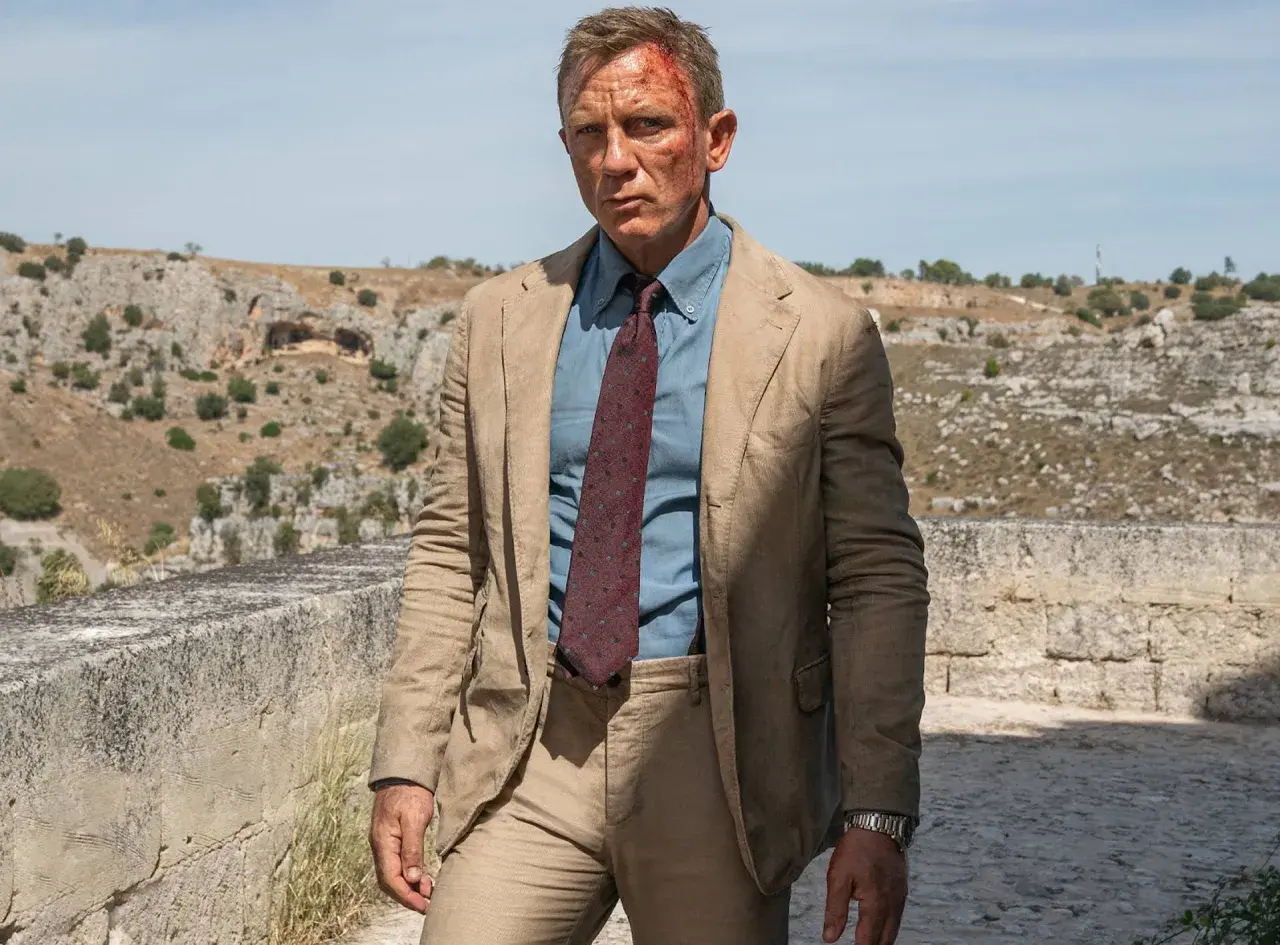 Daniel Craig in einem hellbraunen Anzug, einem blauen Hemd und einer gemusterten Krawatte, typisch für James Bond Outfits, mit leichten Verletzungen im Gesicht.