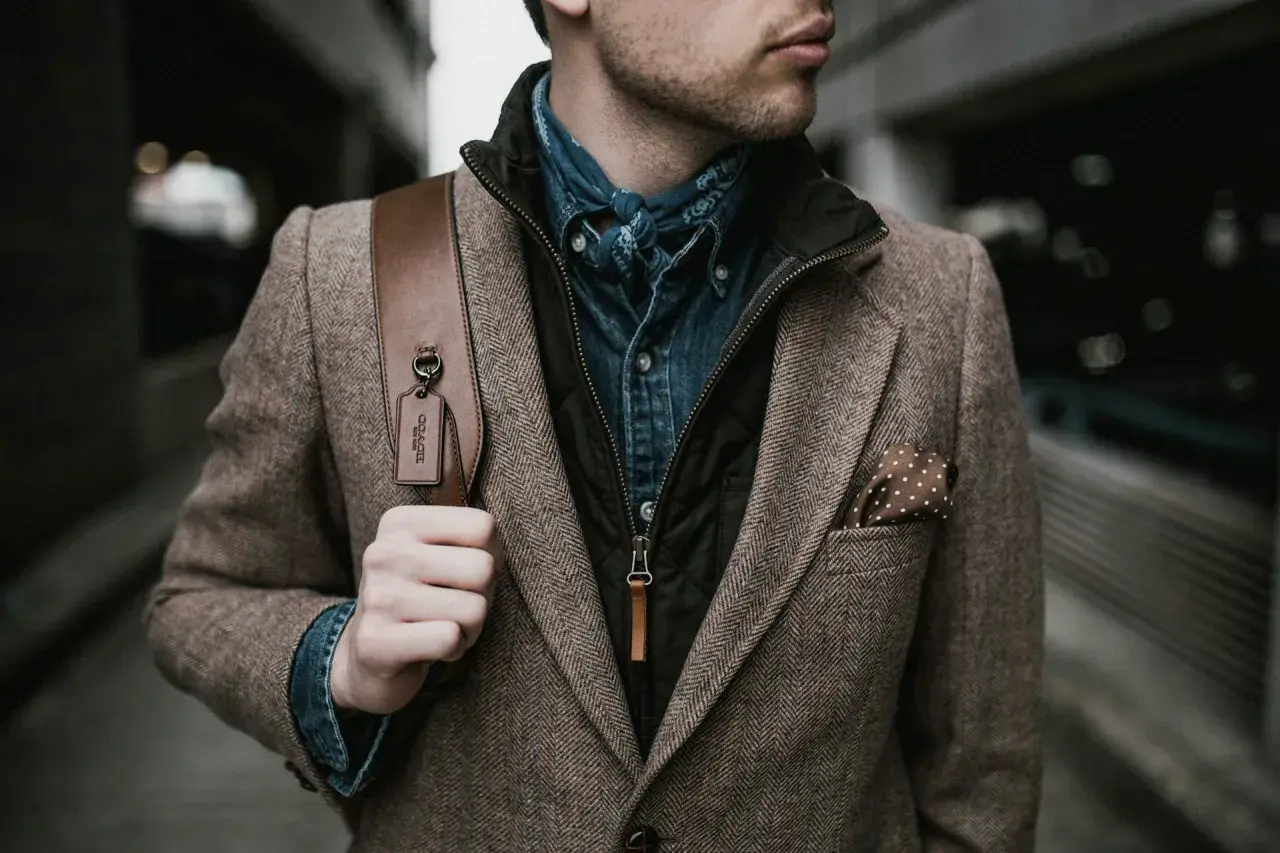 Mann im Tweed-Sakko mit Jeanshemd und Bandana, perfekt für den dresscode chic herren. Braune Ledertasche mit Coach-Anhänger.