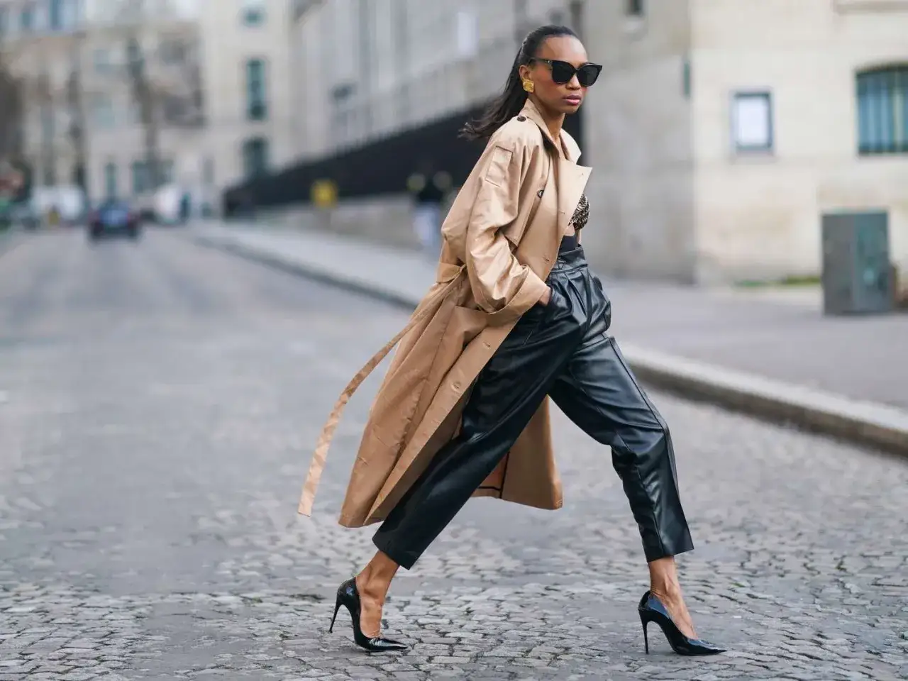 Eine Frau im Trenchcoat und Lederhose, mit hohen Abs&auml;tzen, verk&ouml;rpert pariser chic auf Kopfsteinpflaster.