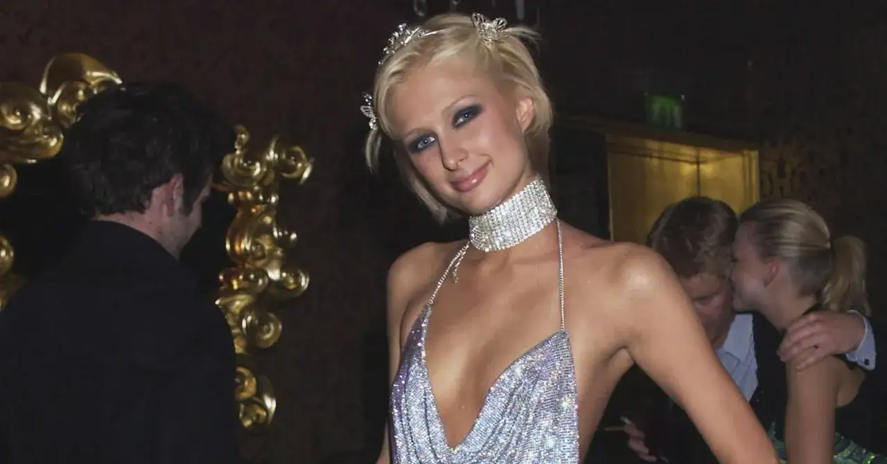 Paris Hilton glänzt in einem funkelnden 2000er Mottoparty-Outfit mit Diadem und Choker. Im Hintergrund tuscheln andere Gäste.