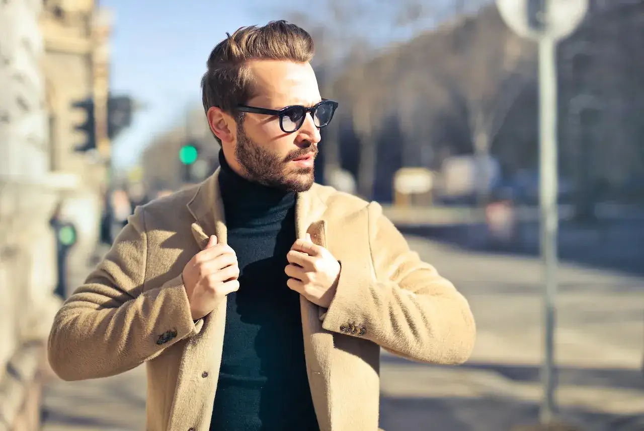 Ein Mann mit Bart und Sonnenbrille, der sich stilvoll kleidet, tr&auml;gt einen beigen Mantel &uuml;ber einem schwarzen Rollkragenpullover.