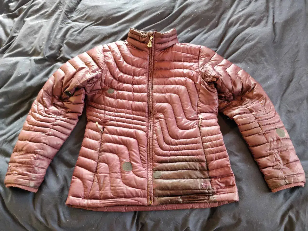 Eine dunkelrote Daunenjacke liegt auf einem dunklen Stoff. Sie ist leicht zerknittert und scheint bereit für die daunenjacke waschen.