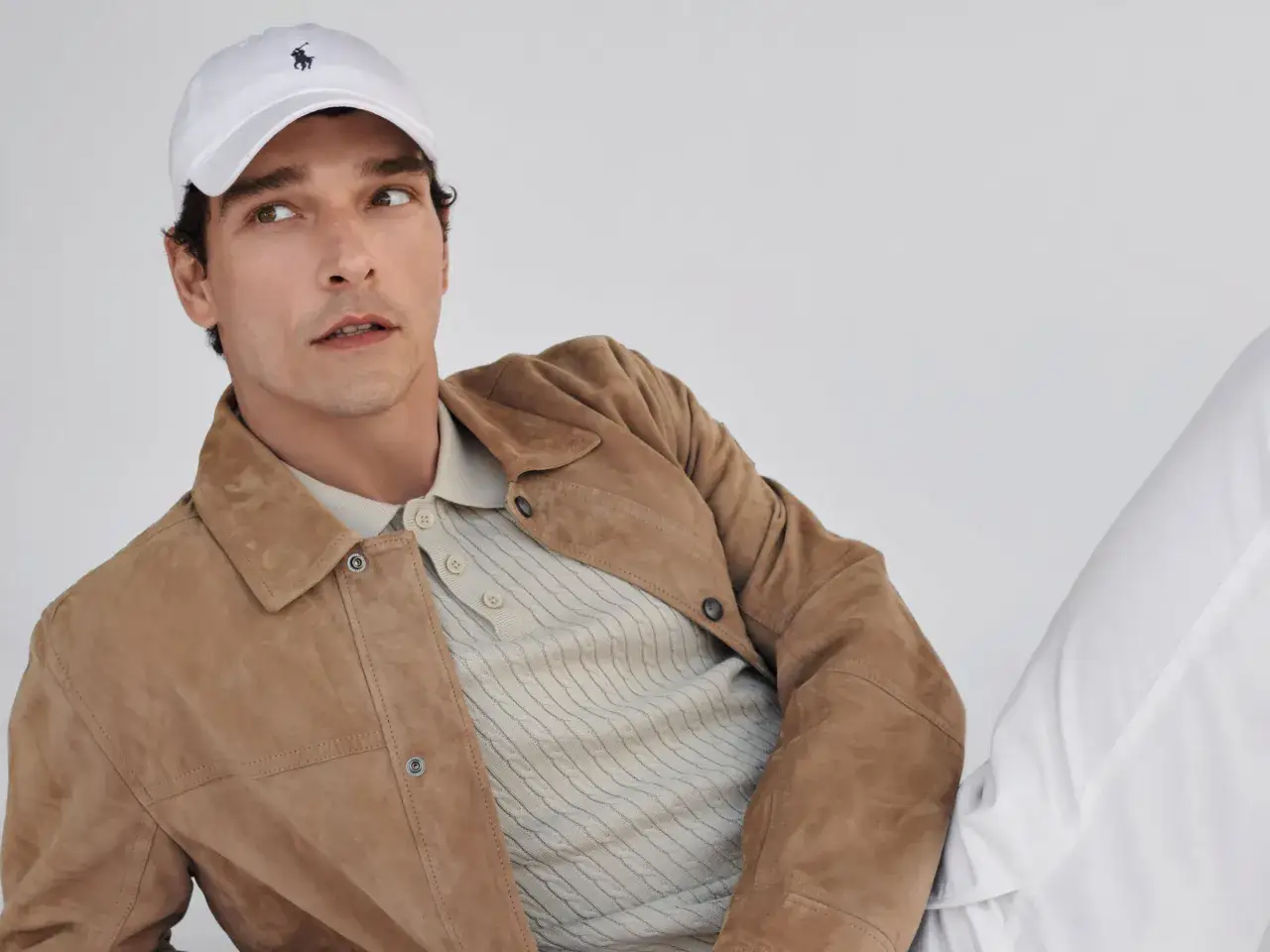 Mann mit brauner Wildlederjacke, beigem Poloshirt und weißer Kappe mit Polo-Logo.
