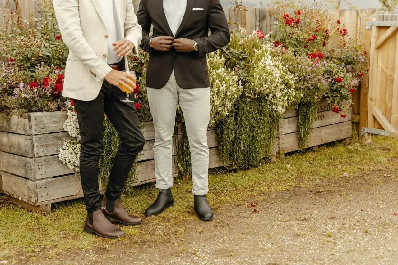 Zwei Männer in Anzügen, einer mit braunen Chelsea Boots, der andere mit schwarzen, stehen vor blühenden Holzkisten.
