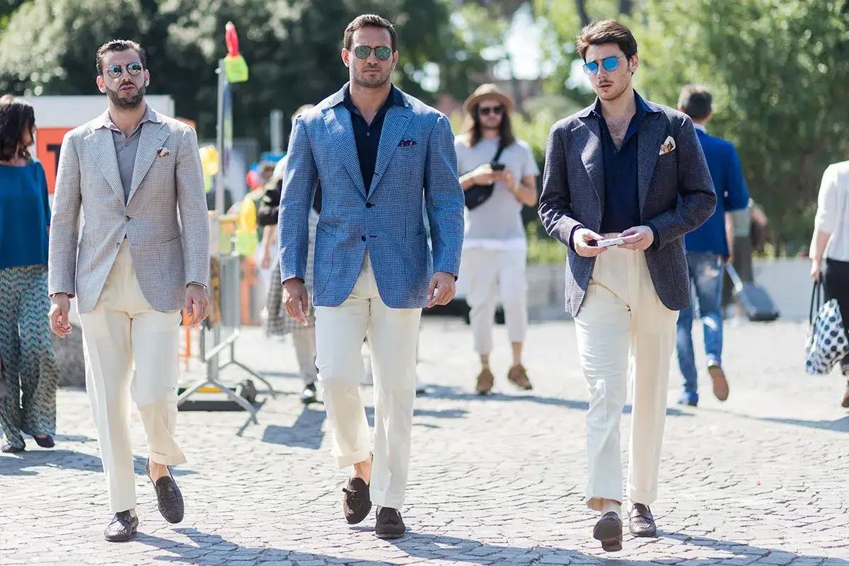 Drei M&auml;nner in eleganten Outfits: Ein Mann tr&auml;gt ein blaues Sakko mit beige Hose, ein anderer ein helles Sakko mit beige Hose.