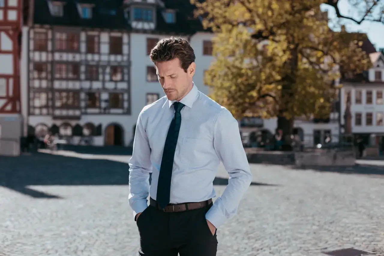 Mann in einem hellblauen Hemd und einer dunkelblauen Krawatte mit Muster steht mit den Händen in den Hosentaschen vor historischer Architektur.