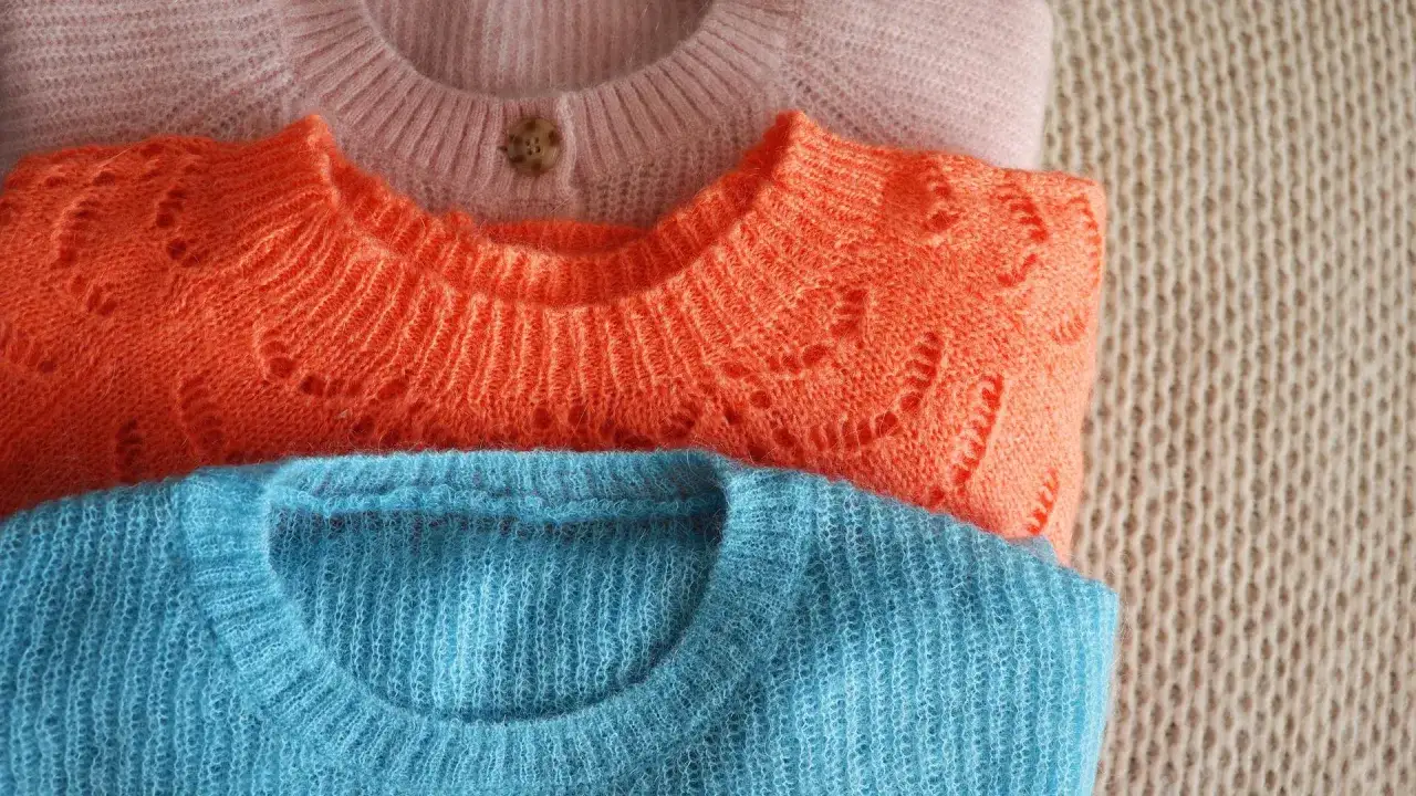 Drei gestapelte Wollpullover in Rosa, Orange und Blau. Der orangefarbene Pullover hat ein schönes Lochmuster. Zeit, den wollpullover waschen.